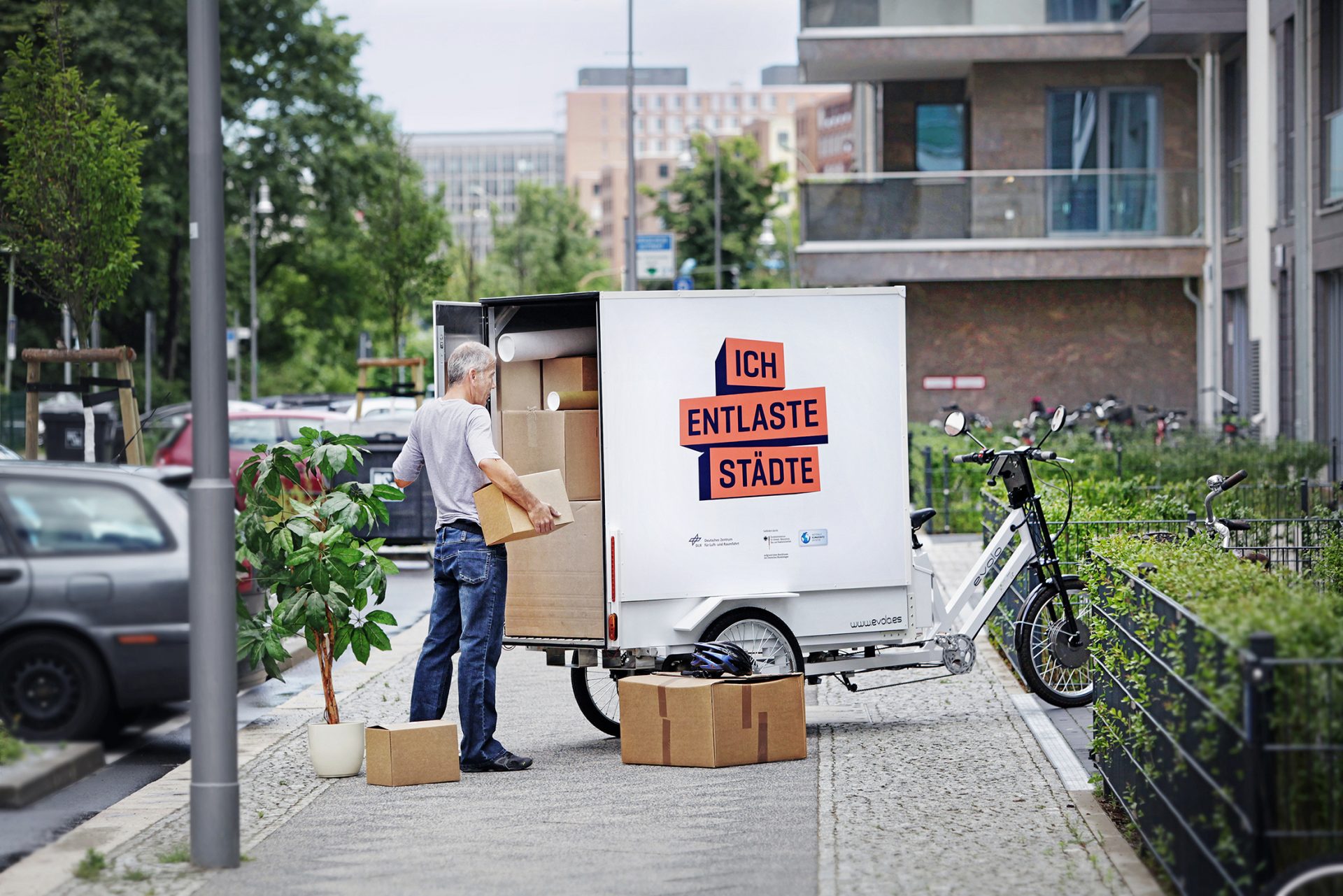 Un hombre carga en la acera una gran bicicleta de carga con cajas de mudanza. En la bicicleta está 