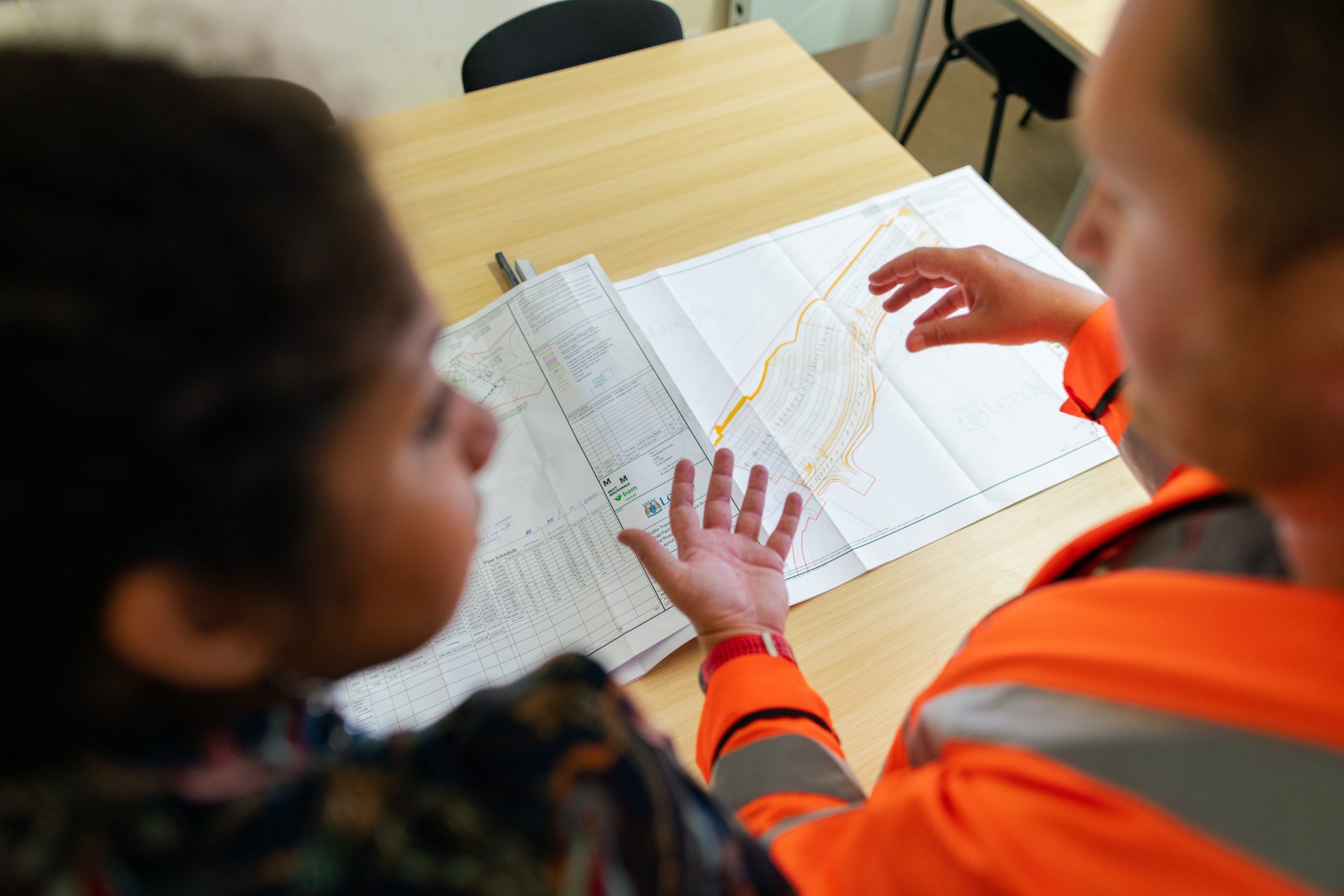 Dos personas están de pie delante de un mapa de planificación sobre la mesa. Una de las personas lleva un chaleco naranja de alta visibilidad.